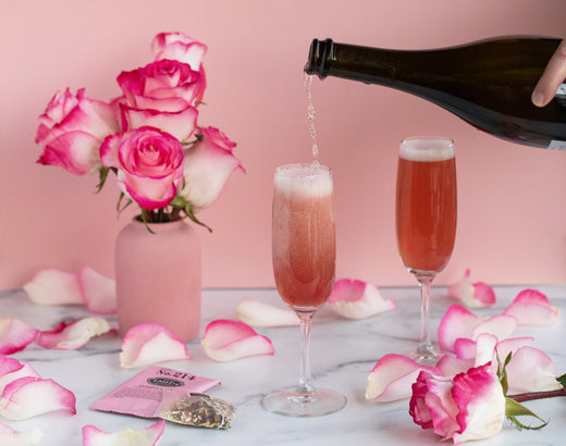 Bottle of prosecco pouring into champagne glass filled with Chivalry cocktail with roses surrounding the glasses.