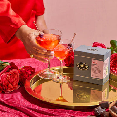 Hand holding coupe glass filled with Sands of Time cocktail with large ice cube on a gold tray with grey box of Kissmet tea.