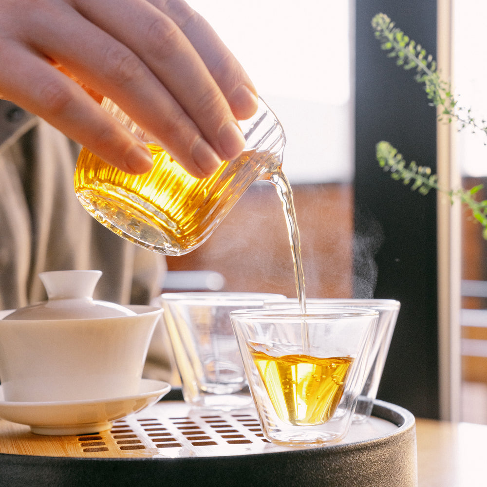 Brewed tea being poured from a glass decanter into a glass gaiwan cup on a gaiwan tray.