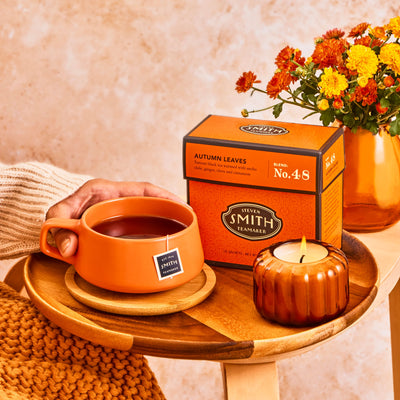 Deep orange box of Autumn Leaves on a wooden tray alongside a lit candle and an orange teacup with a sachet in it.