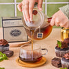 Person pouring tea from teacup into mug with box of Chocolate Peppermint Pu-erh in background, surrounded by chocolate cupcakes.