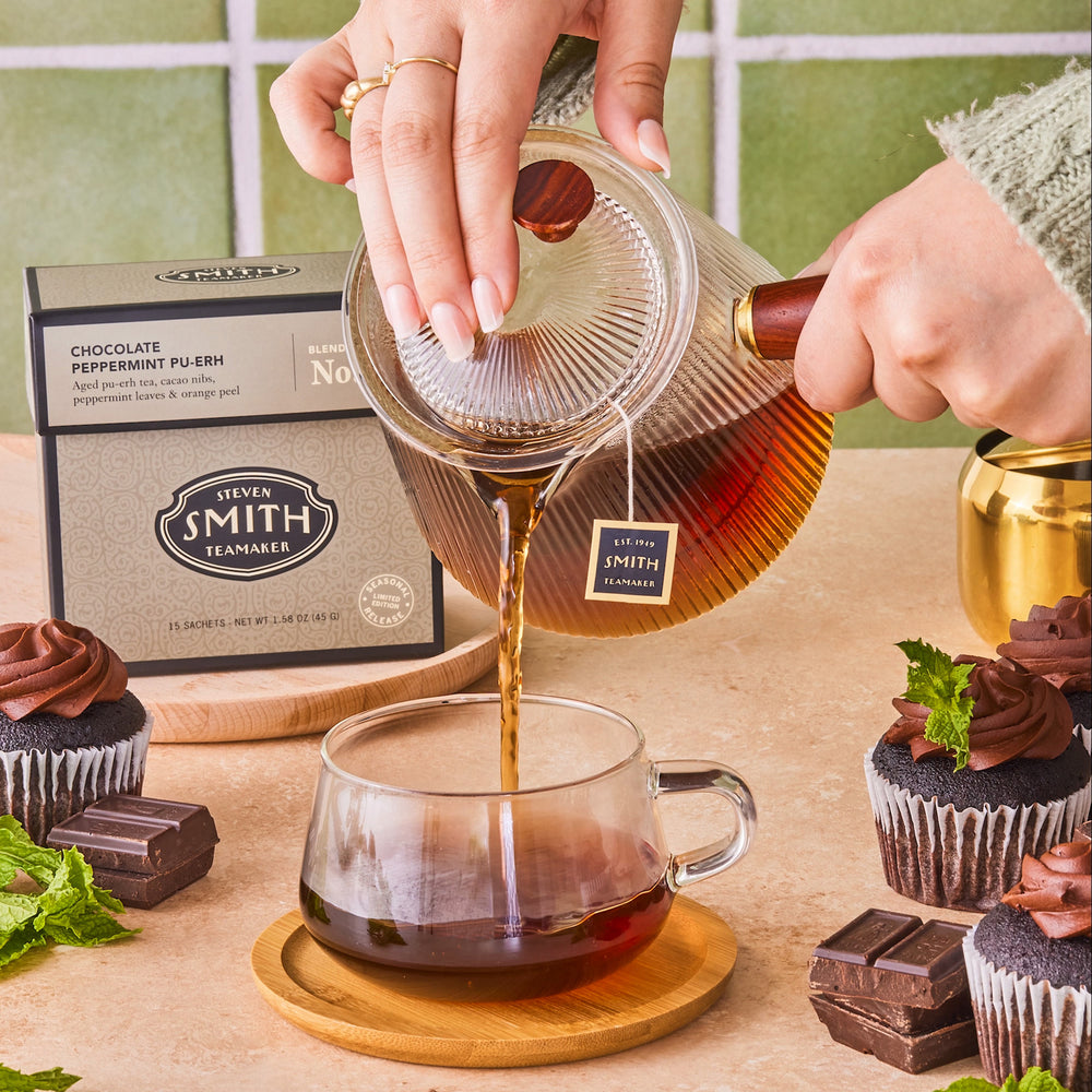 Person pouring tea from teacup into mug with box of Chocolate Peppermint Pu-erh in background, surrounded by chocolate cupcakes.