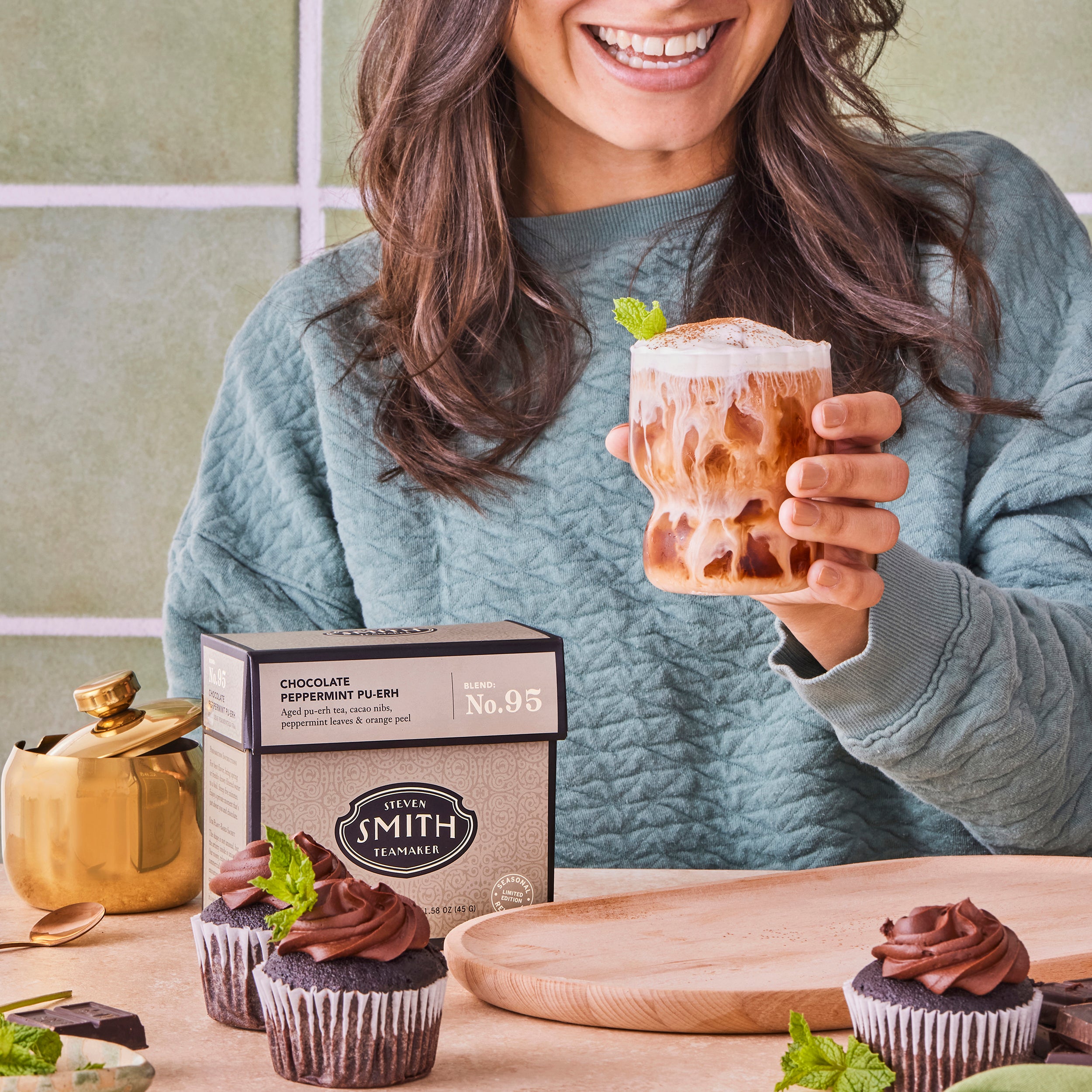 Woman holding a glass of iced Chocolate Peppermint Pu-erh with cold foam at a table with box of tea and cupcakes.