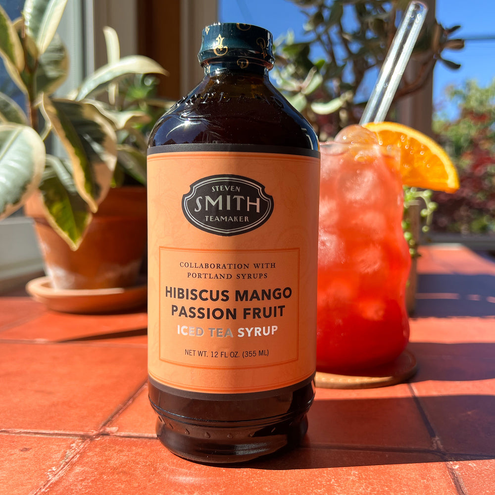 Bottle of Hibiscus Mango Passion Fruit Syrup with an orange label with mocktail in background on counter.