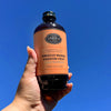 Bottle of Hibiscus Mango Passion Fruit Syrup with an orange label held in the air with sky in the background.