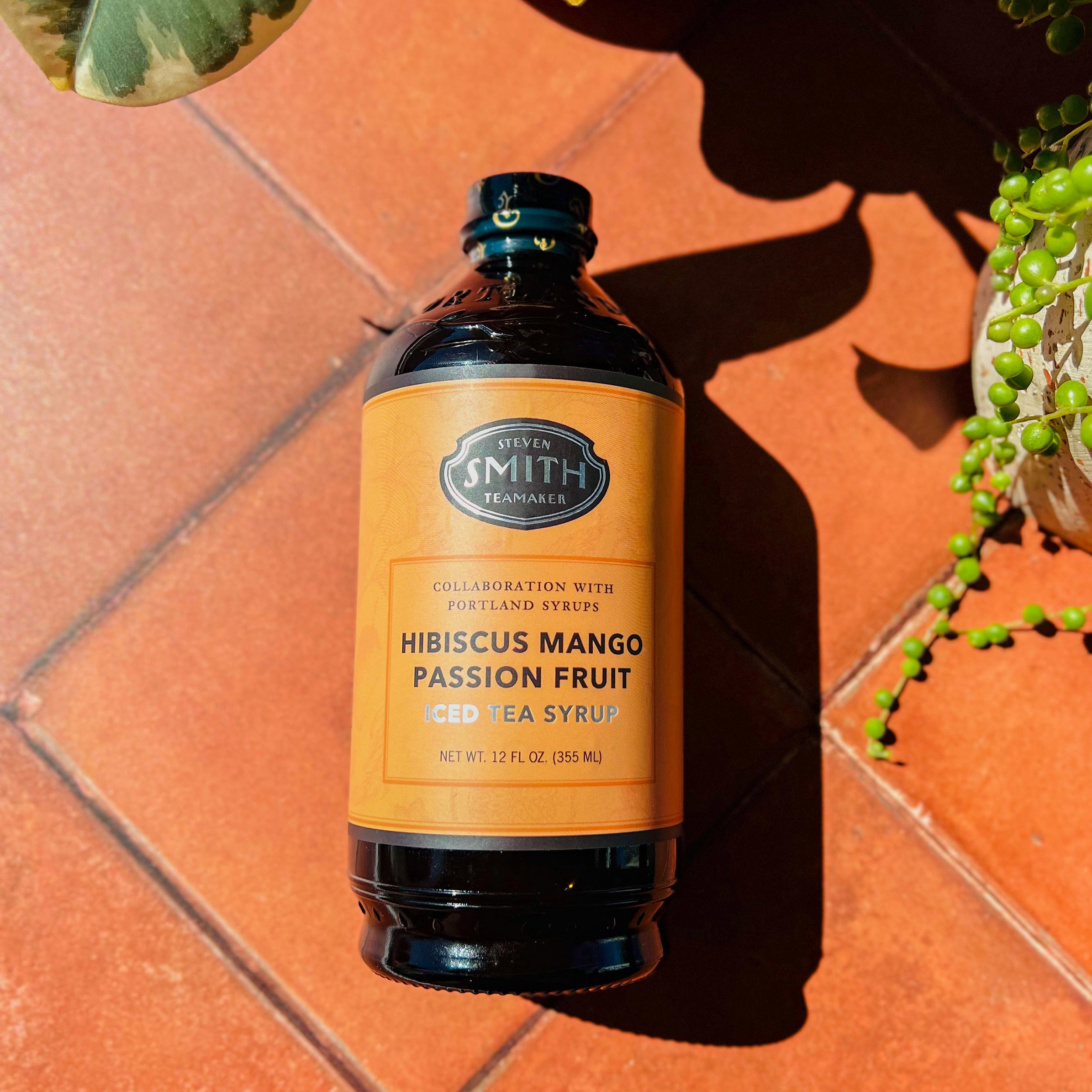 Bottle of Hibiscus Mango Passion Fruit Syrup with an orange label on red tile background.