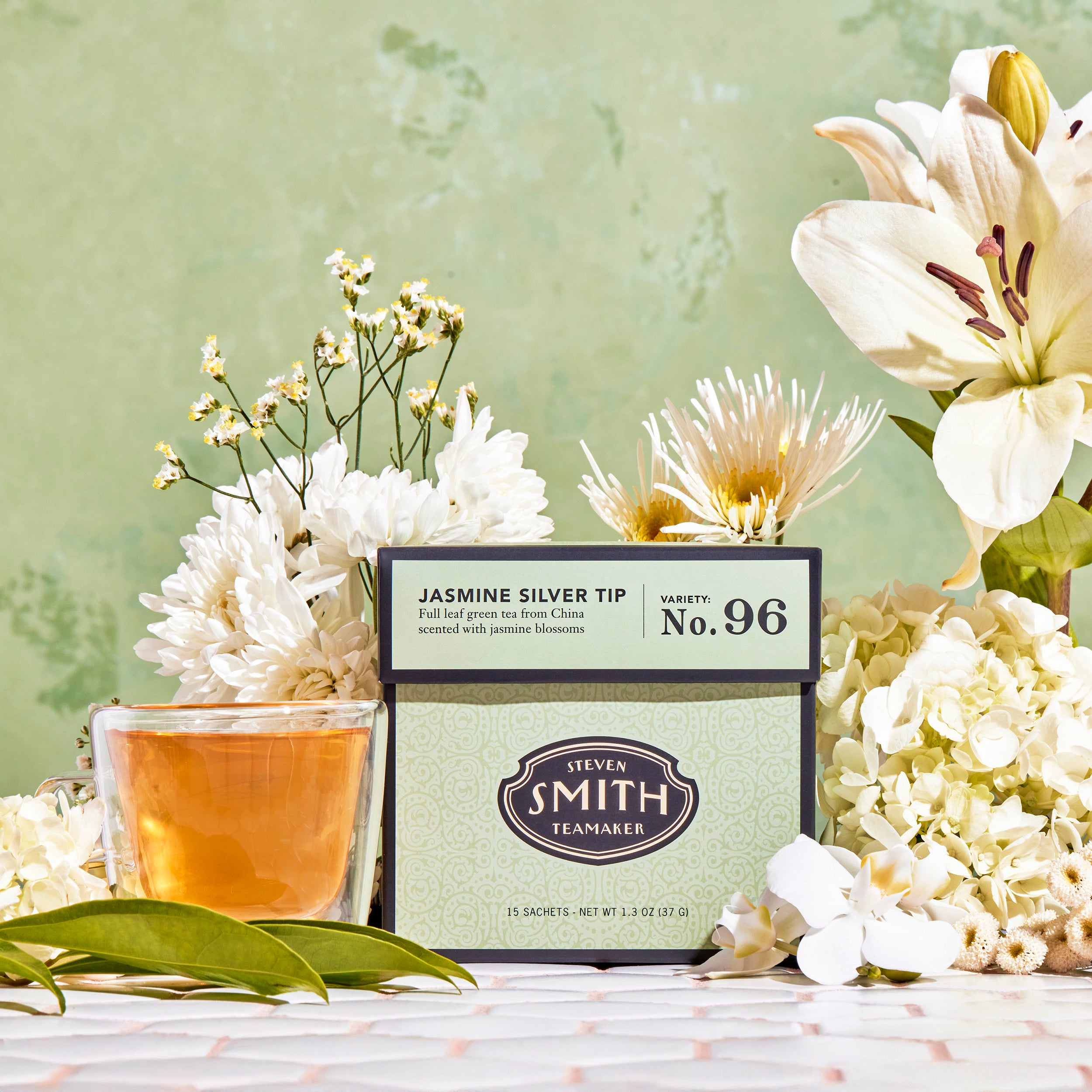 Green box of Jasmine Silver Tip against a pale green background surrounded by white flowers beside a cup of brewed tea.