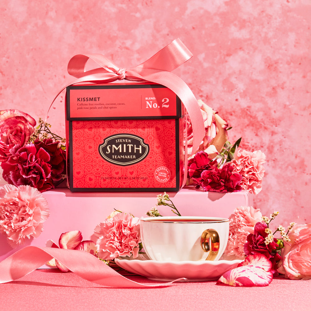 Deep pink colored box of Kissmet tea surrounded by flowers and a teacup.