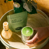 Hand holding a glass cup with matcha latte beside a green pouch of Matcha Latte mix and a matcha whisk.