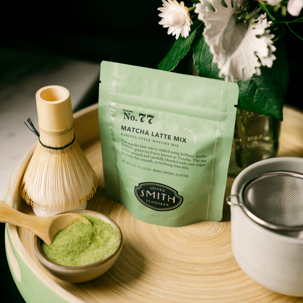 Green pouch of Matcha Latte Mix on a wooden tray with a matcha whisk, bowl of loose Matcha Latte mix and a matcha bowl.