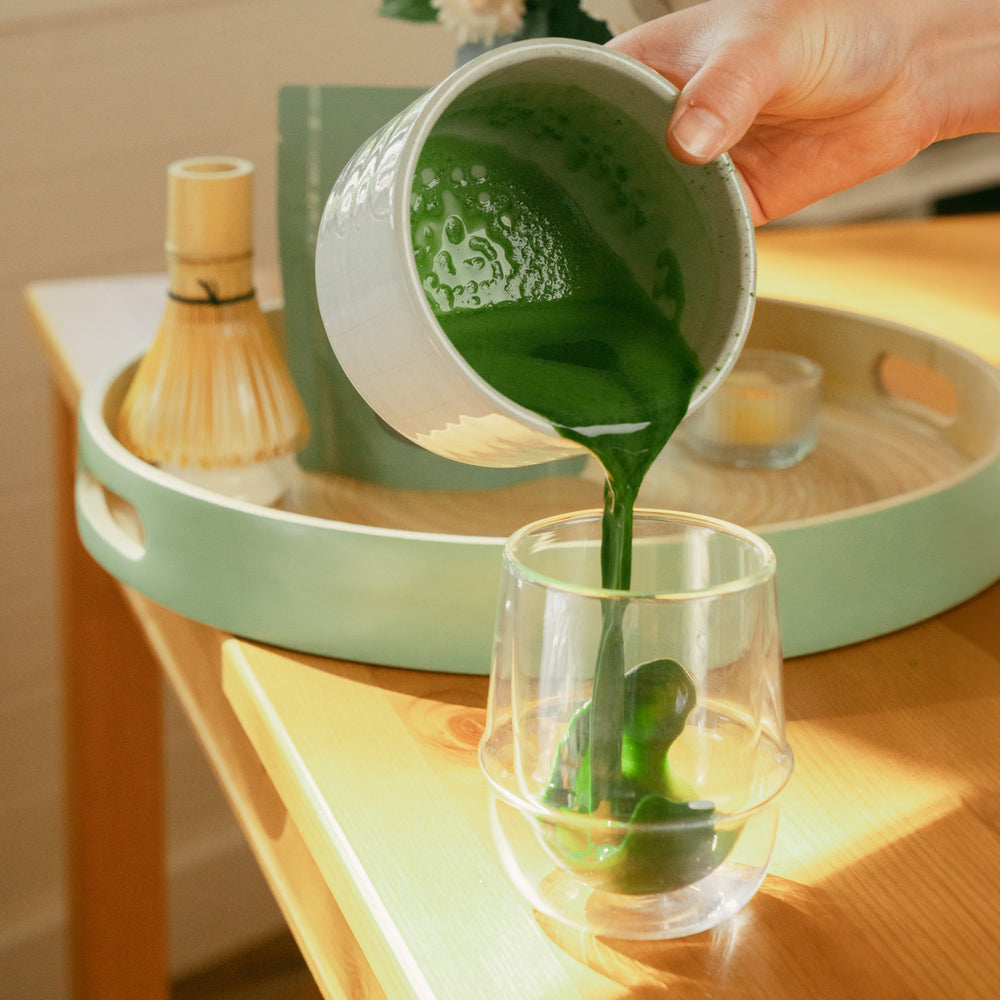 Frothed matcha being poured from a matcha bowl into a glass cup in a kitchen setting.