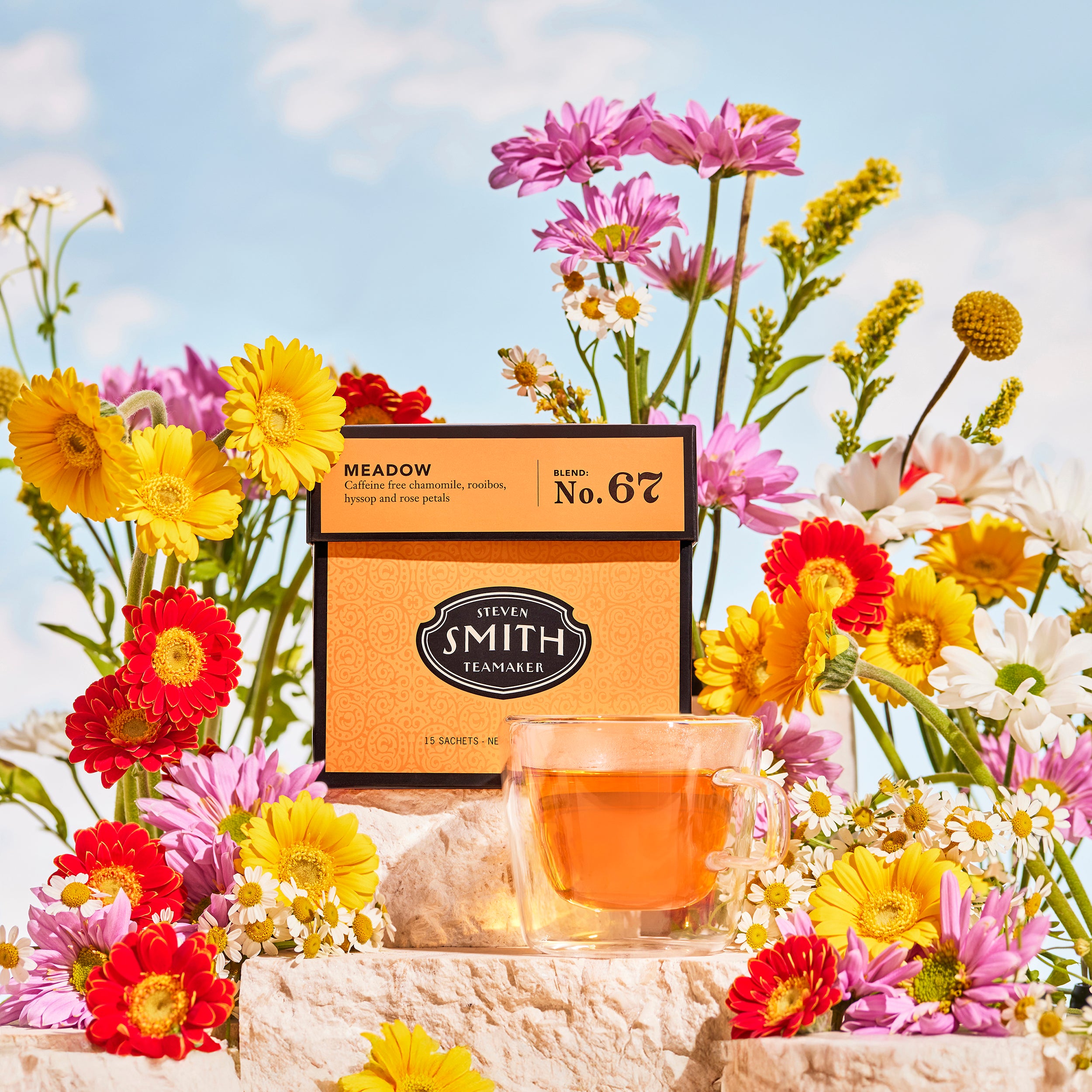 Orange box of Meadow surrounded by brightly colored flowers against a blue sky beside a cup of brewed tea.