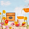 Orange box of Meadow surrounded by brightly colored flowers against a blue sky beside a cup being filled with brewed tea poured from a glass teapot.