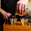 Golden brewed Nan Nuo Shan Pu-erh being poured from a glass decanter into two glass Gaiwan cups on a wooden Gaiwan tray.