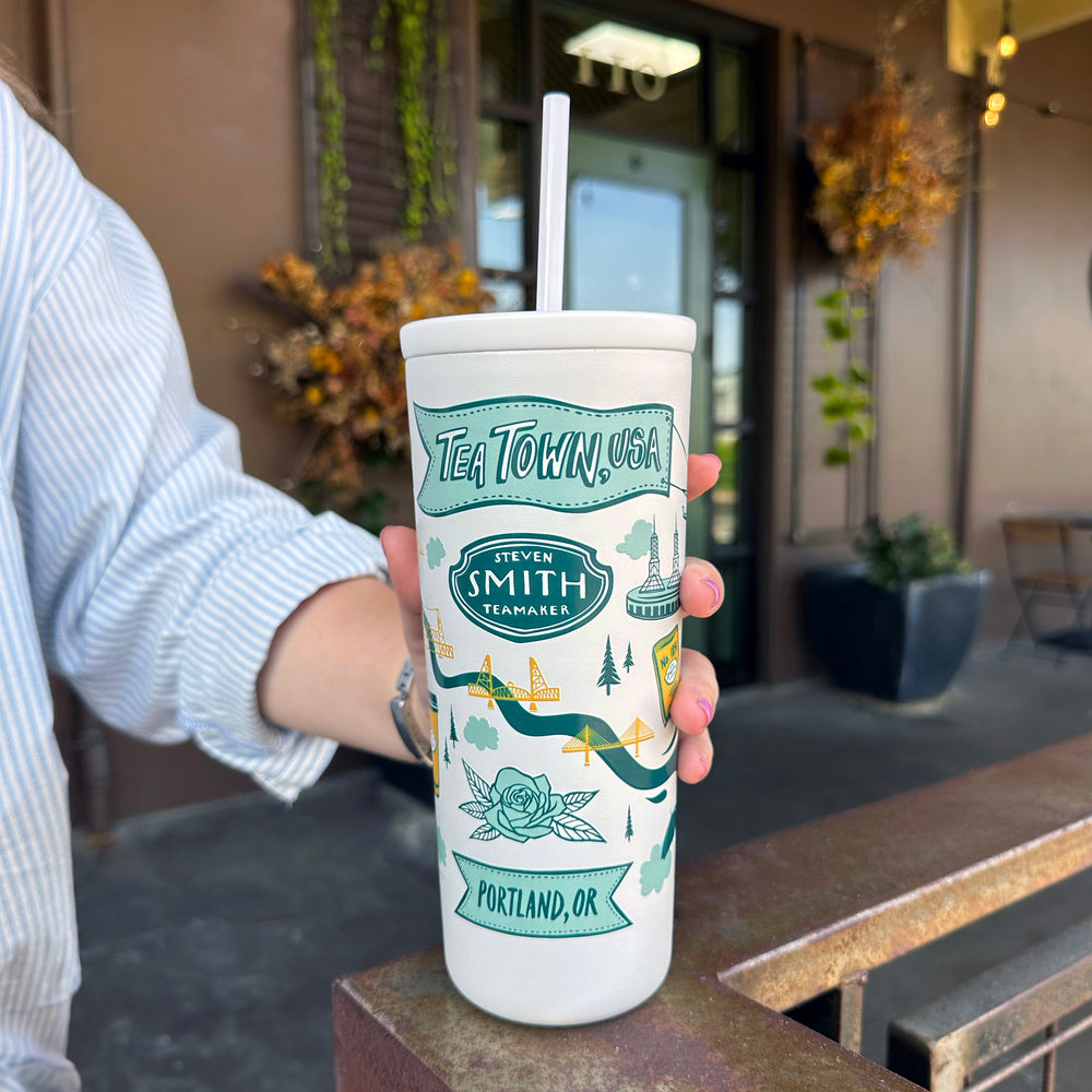 White cold cup with straw and Tea Town, USA design in blue and yellow held in front of Smith Teamaker HQ.