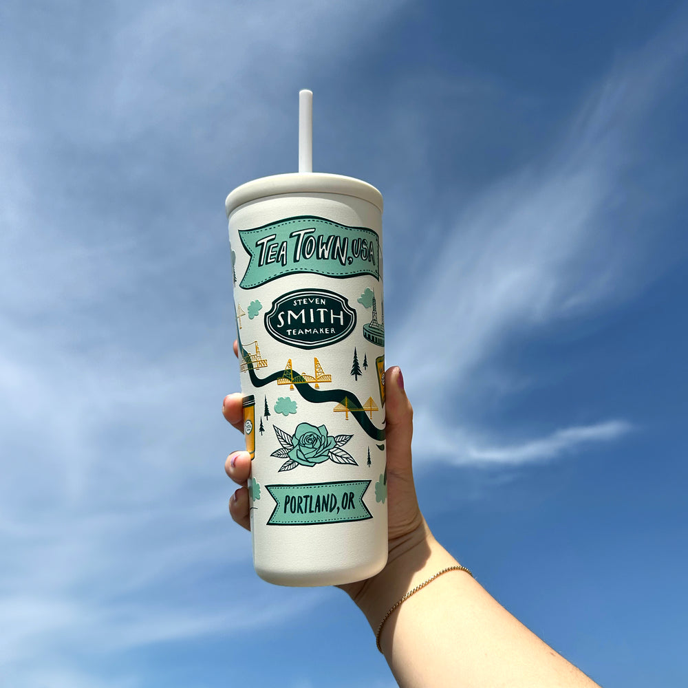 White cold cup with straw and Tea Town, USA design in blue and yellow held in front of a blue sky.