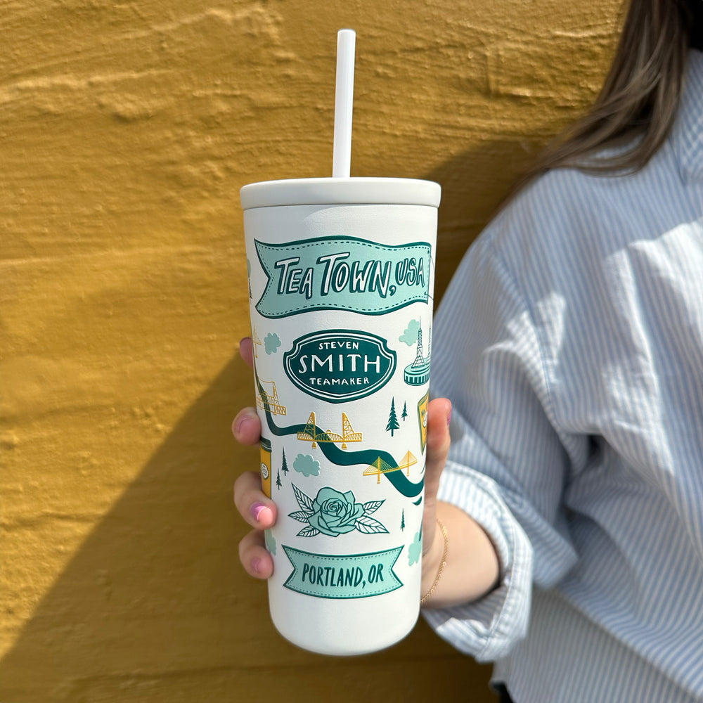 White cold cup with straw and Tea Town, USA design in blue and yellow held in front of a yellow wall.